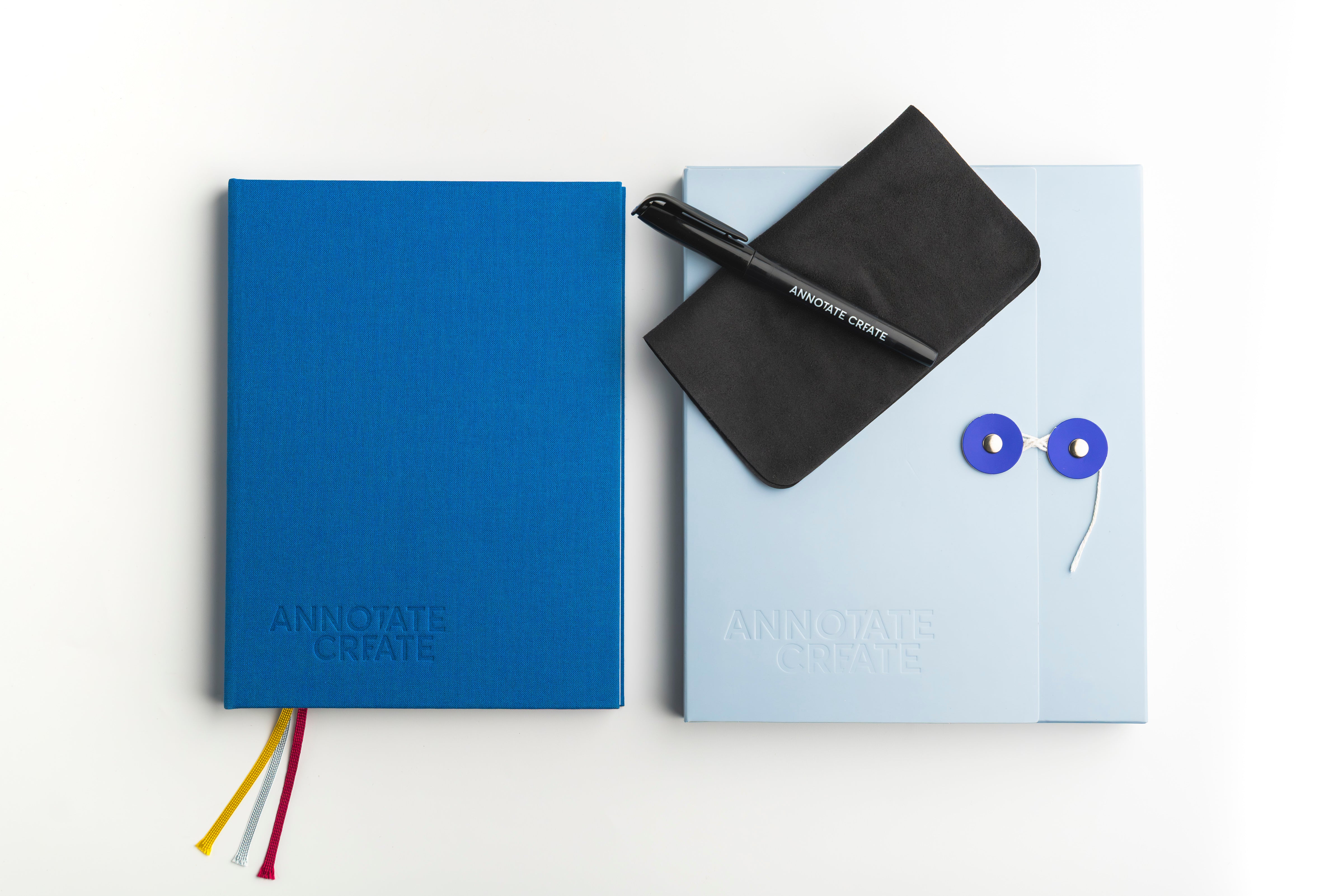 Two Annotate Create notebooks on a white background, one in textured blue with multicolor ribbon bookmarks and one in pale blue with a black cover slip and pen.