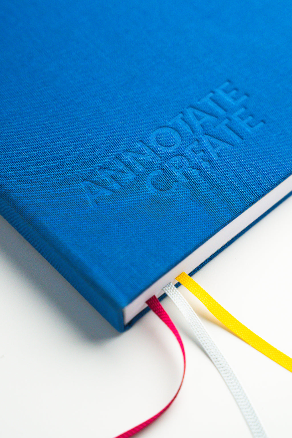 Close-up of Nōto notebook's blue linen fabric cover with embossed logo and spine showing red, white, and yellow ribbon bookmarks