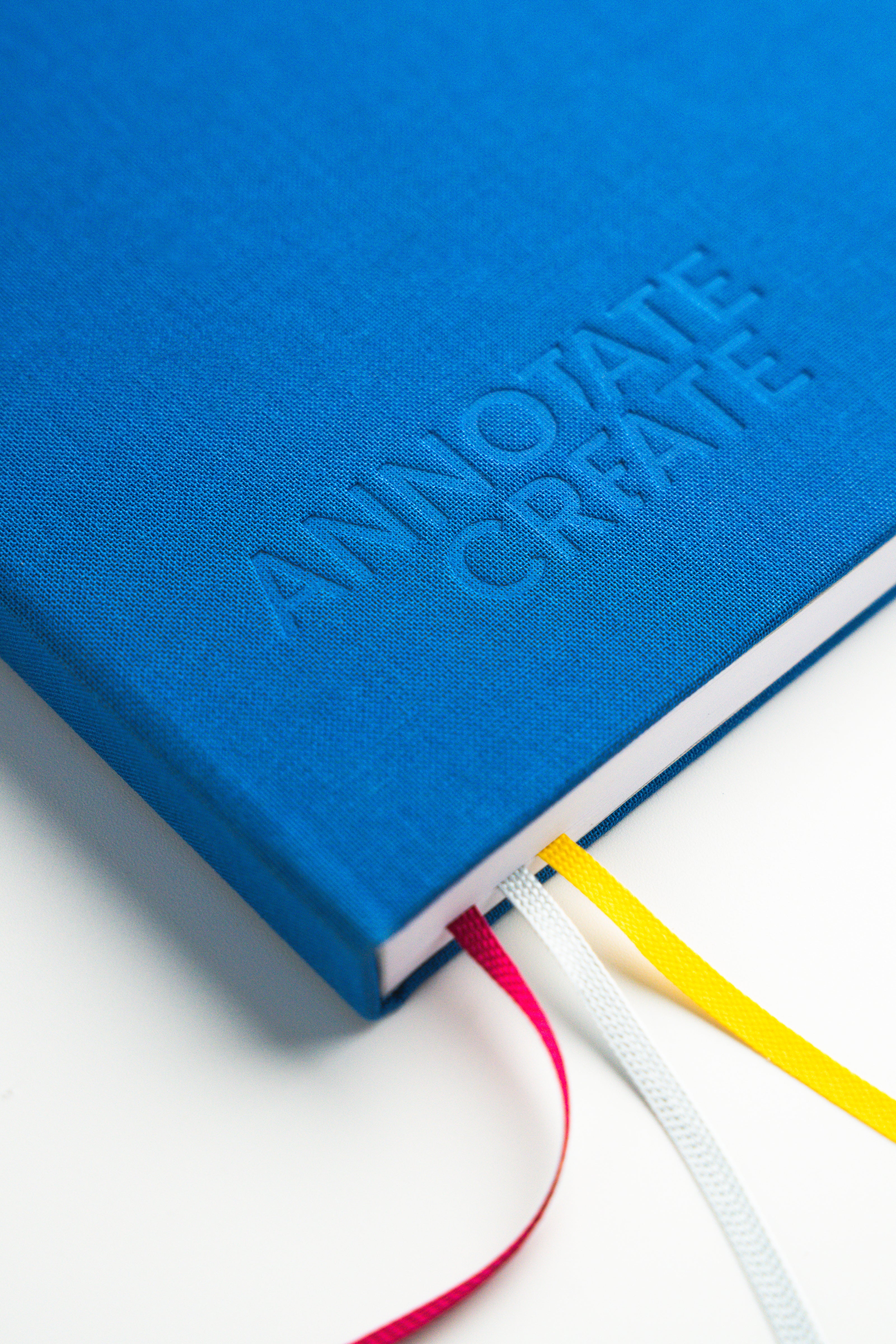 Close-up of Nōto notebook's blue linen fabric cover with embossed logo and spine showing red, white, and yellow ribbon bookmarks